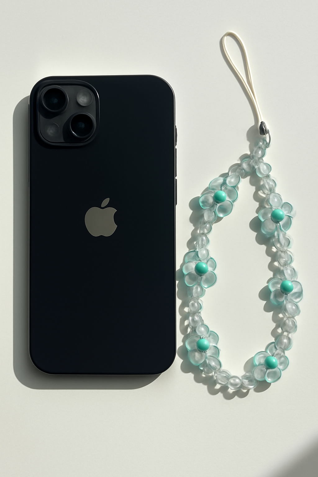 A clear and aqua blue beaded floral phone lanyard positioned next to a black smartphone on a stark white background with harsh shadows.